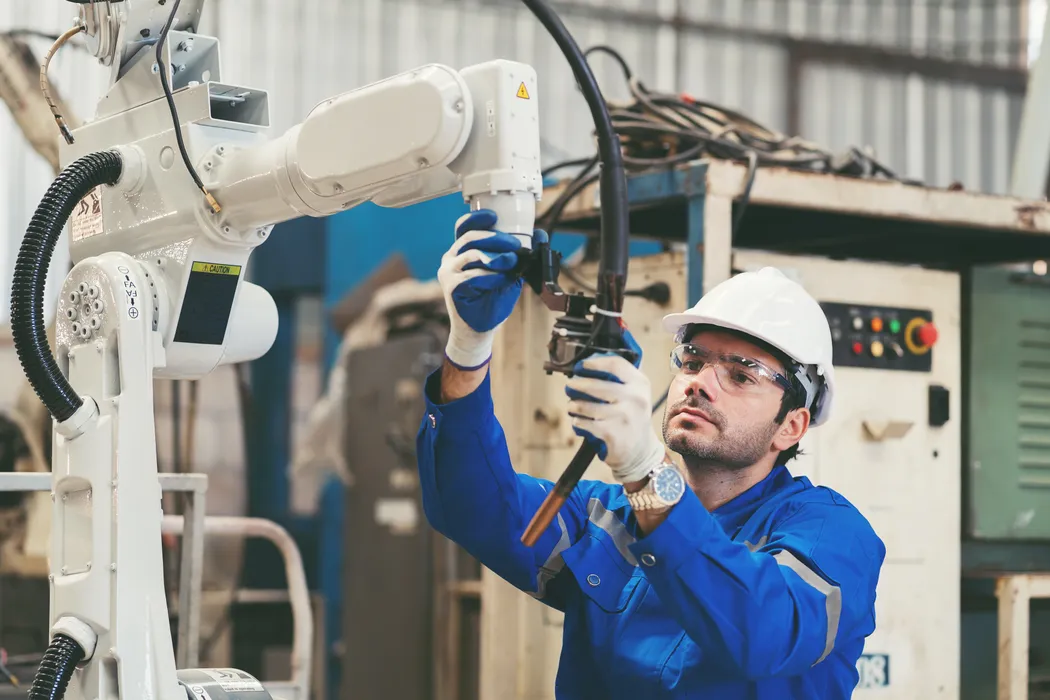 Arbeiter in blauer Uniform und weißem Schutzhelm justiert in einer Industrieanlage einen Roboterarm.