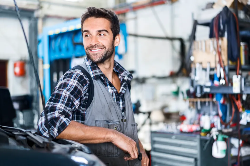 Handwerker in Arbeitskleidung lehnt in einer Werkstatt an der Motorhaube eines Autos. 